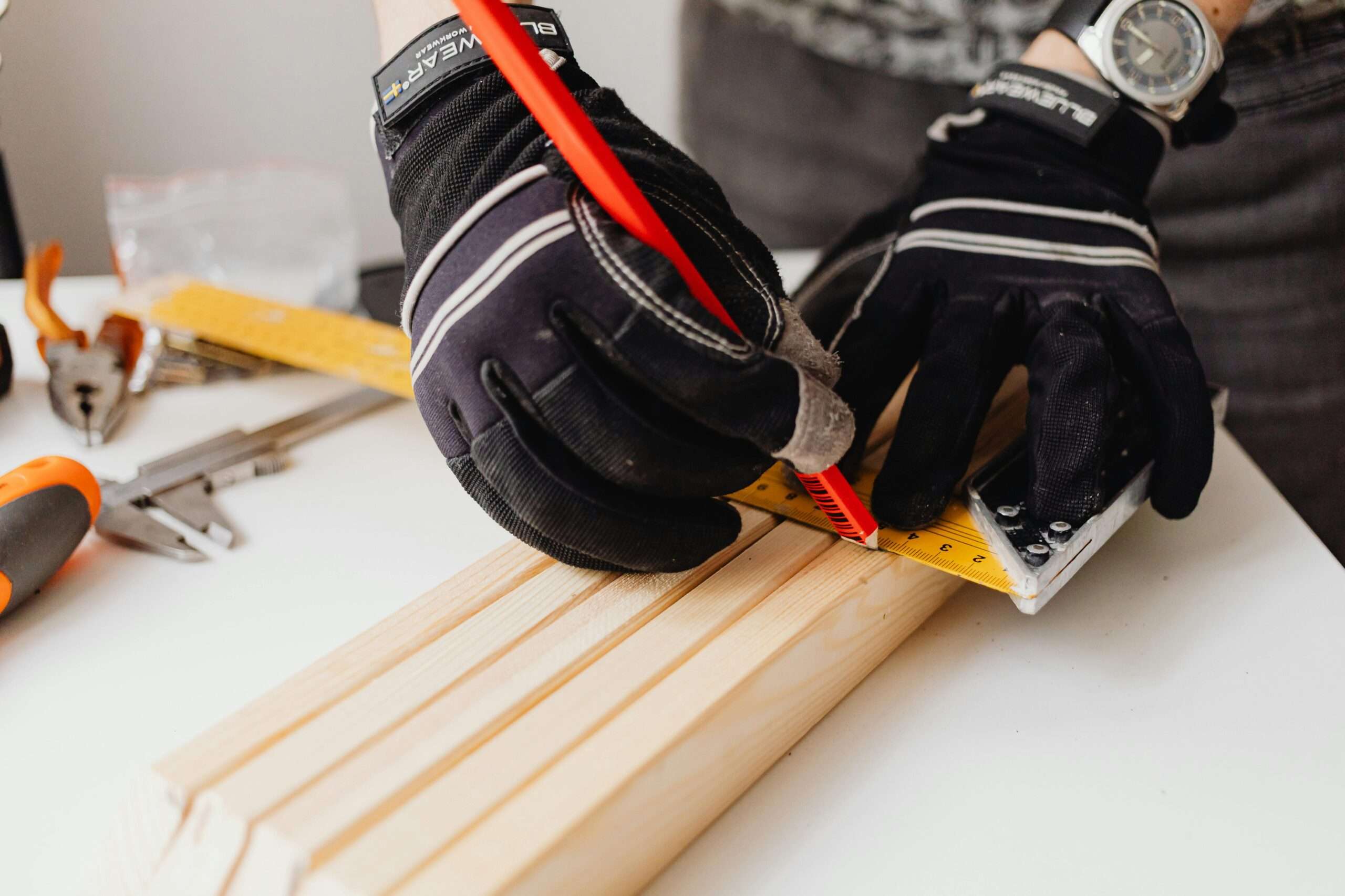 Close-up of gloved hands using ruler to measure wood in a carpentry workshop. Tools are visible in the background.